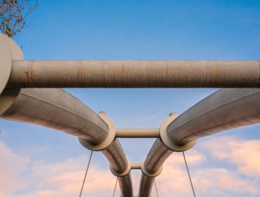 Symmetrical steel pipes supporting a suspension bridge over a river.